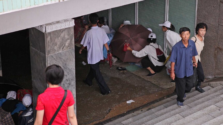 Looking down into the entrance of an underpass people walk up steps, and people shield others with umbrellas.