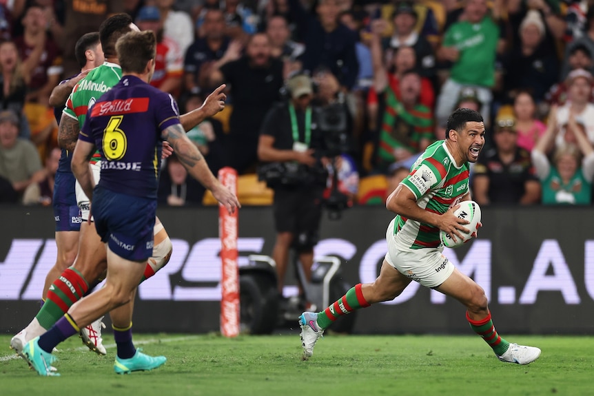 South Sydney NRL player Cody Walker grins as he races in to score a try while other players trail behind him.