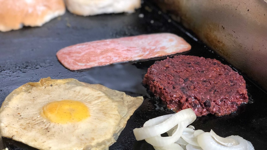 A fake egg, meat patty and bacon rasher on a frying pan.