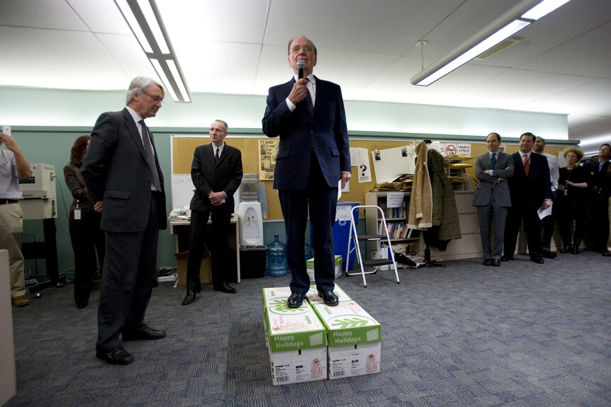 Murdoch stands on two boxes and gives a speech in the middle of the office.