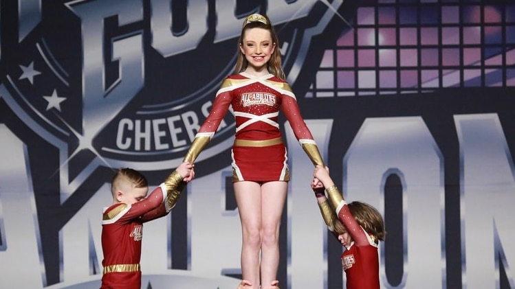 A girl in a red cheerleading outfit with a big smile on her face is being held up by a group of cheerleaders