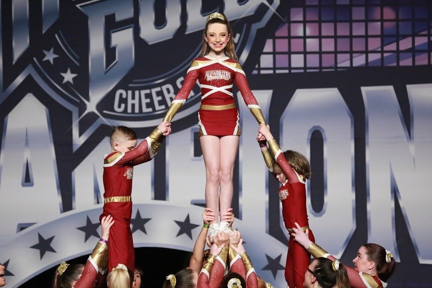 A girl in a red cheerleading outfit with a big smile on her face is being held up by a group of cheerleaders