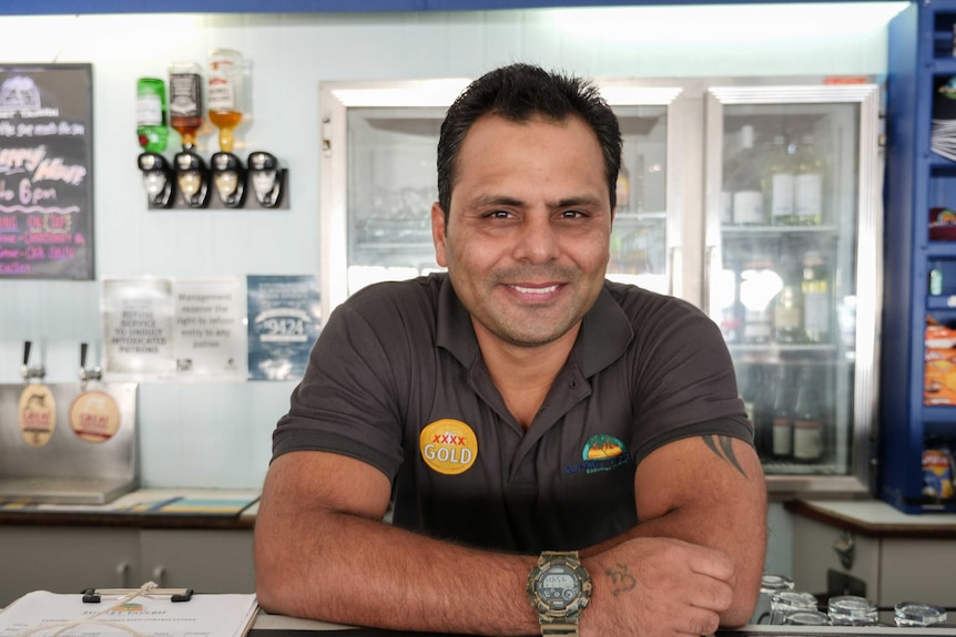 An image of a middle aged man with dark features leaning on a bar. He is smiling at the camera.