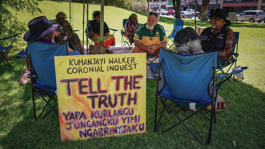 A dozen Indigenous Australians sitting in lawn chairs on a lawn in a town.