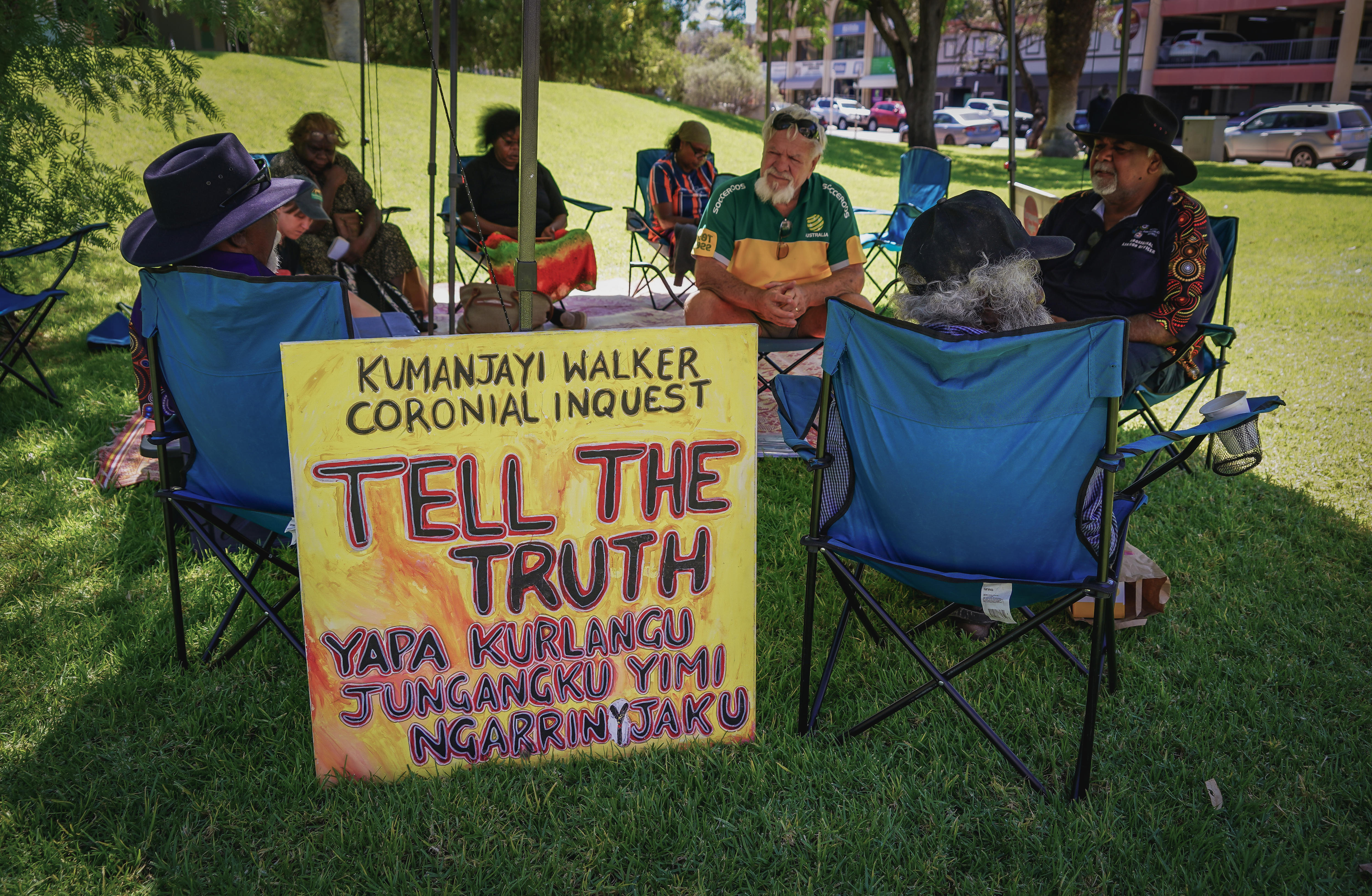 十几名澳大利亚原住民坐在草坪椅上接受法律在一个镇上。” class=