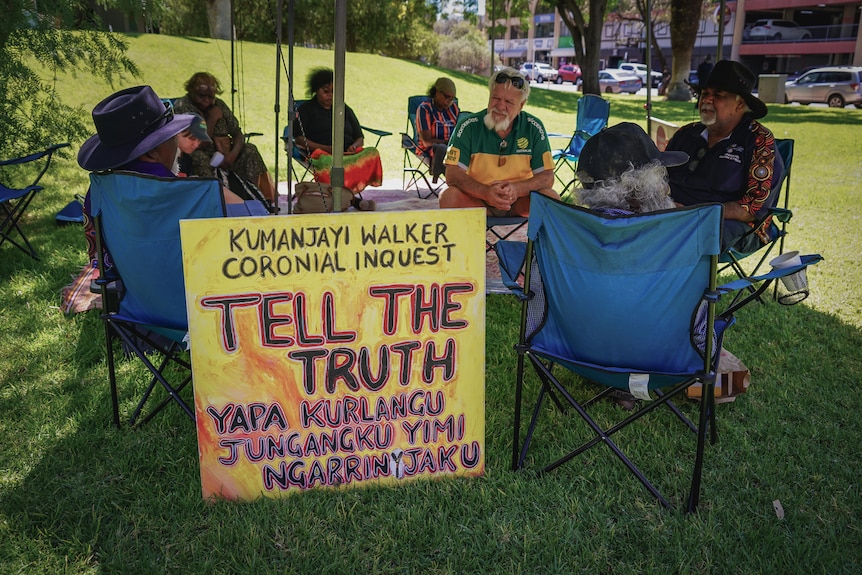 A dozen Indigenous Australians sitting in lawn chairs on a lawn in a town.