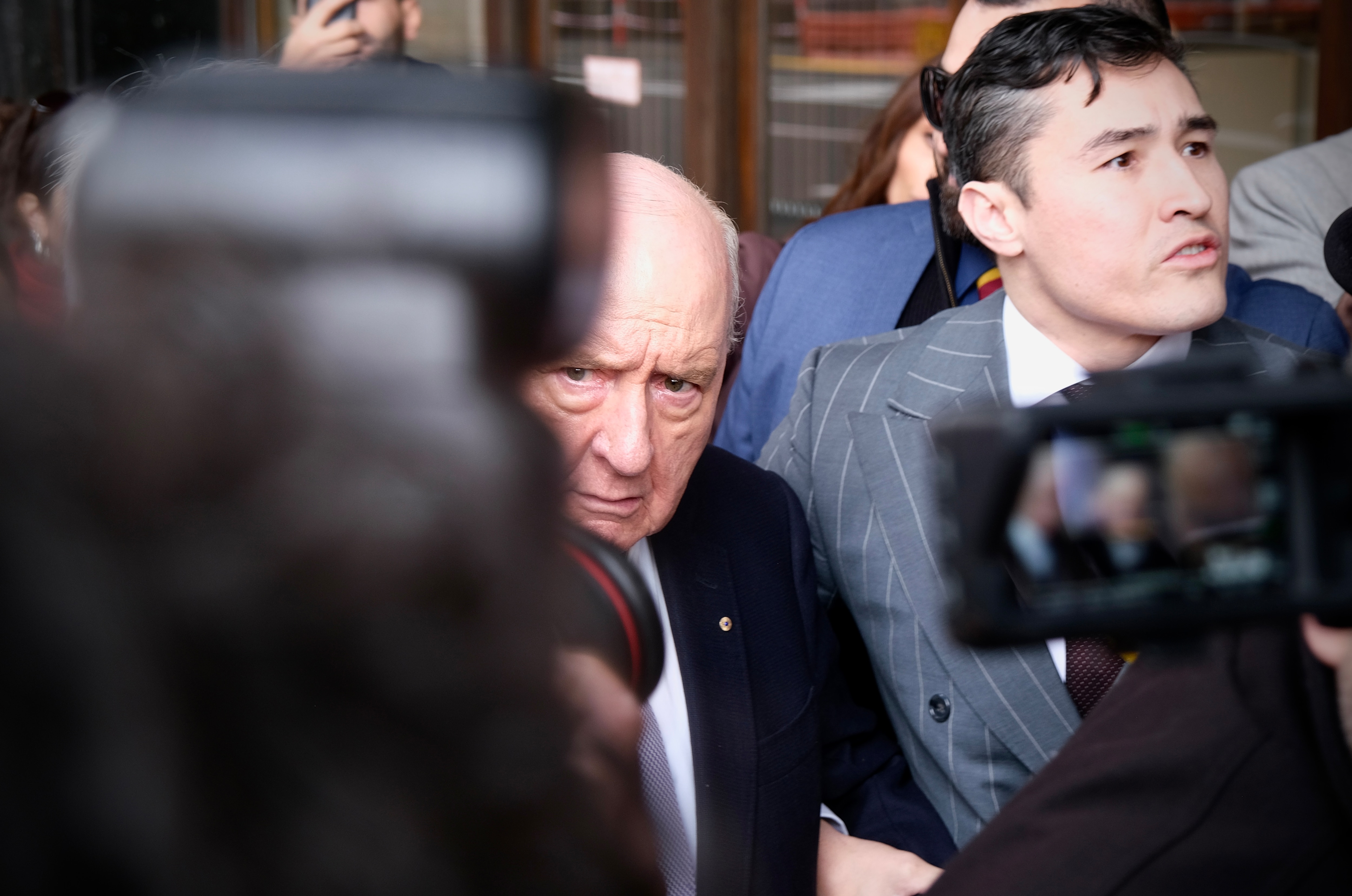 Alan Jones is an elderly, bald man in the middle of a media scrum.