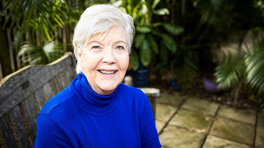 Woman in blue jumper with white hair sits in garden on wooden chair, looking at camera.
