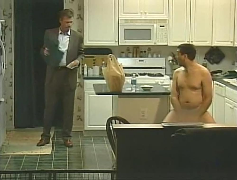 A nude man sits on a kitchen stool while a suited man enters from behind