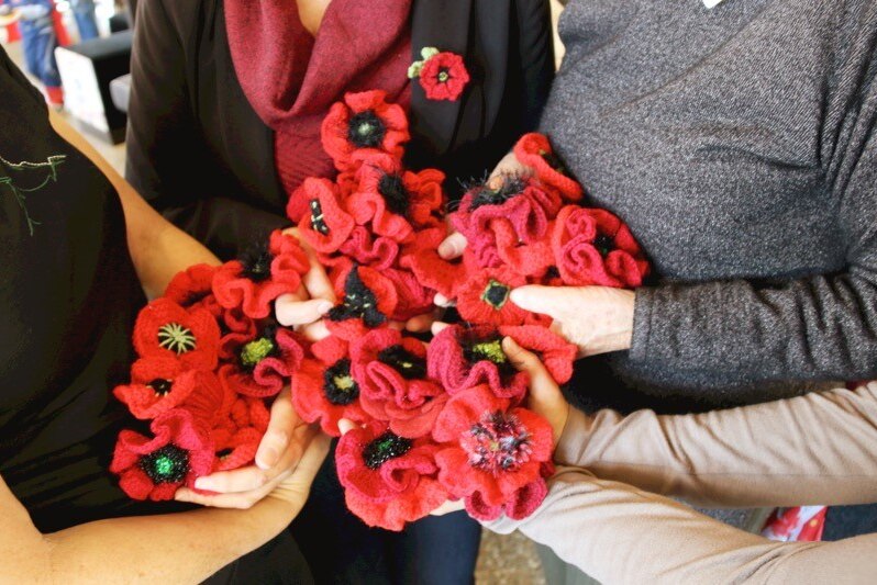 Four women hold in their hands a collection of knitted red poppies.