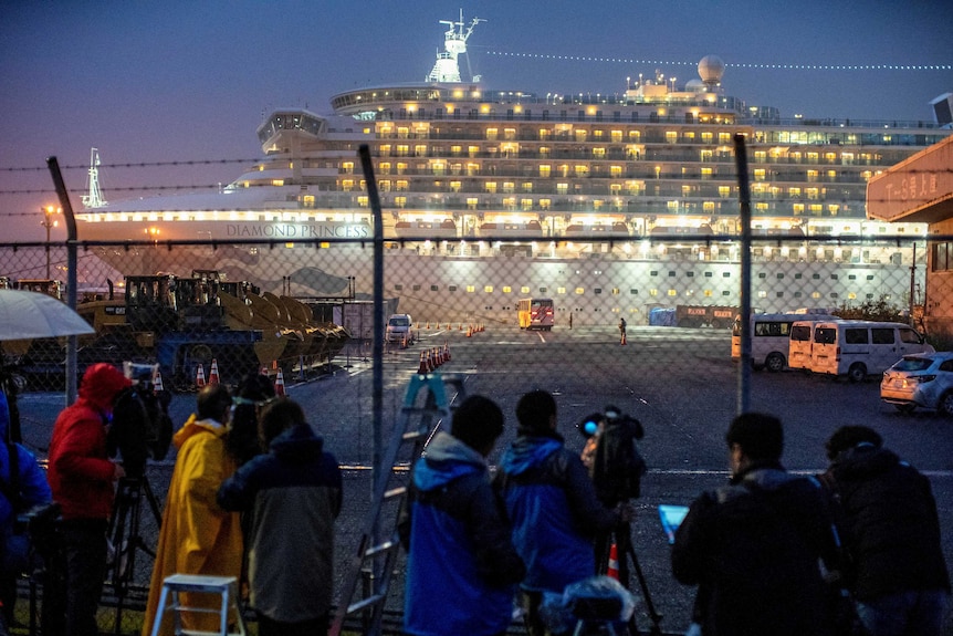 The Diamond Princess lit up at night while media looks through a chain link fence