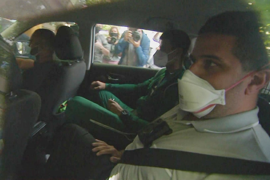 A man in green track pants wearing a mask is sitting with another man as photographers take photo from outside.