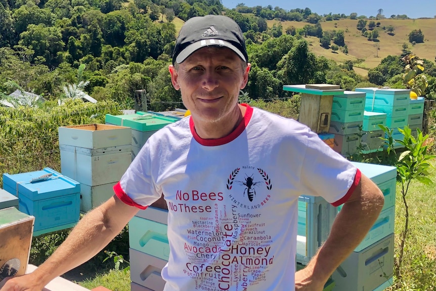 A man poses next to beehives with mountains in the background.