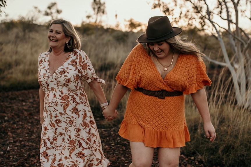 A blonde girl wearing a khaki wide-brimmed hat and short orange dress golds hands with an older woman wearing a white  dr