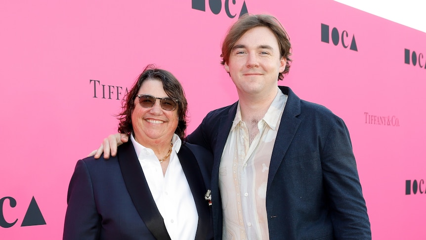 A mother, wearing dark glasses, and son, both wearing suits, pose in front of a pink background