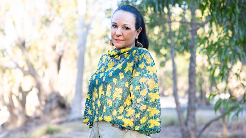 an indigenous woman with a black ponytail wears a top with a green and yellow wattle design, the bush in the background