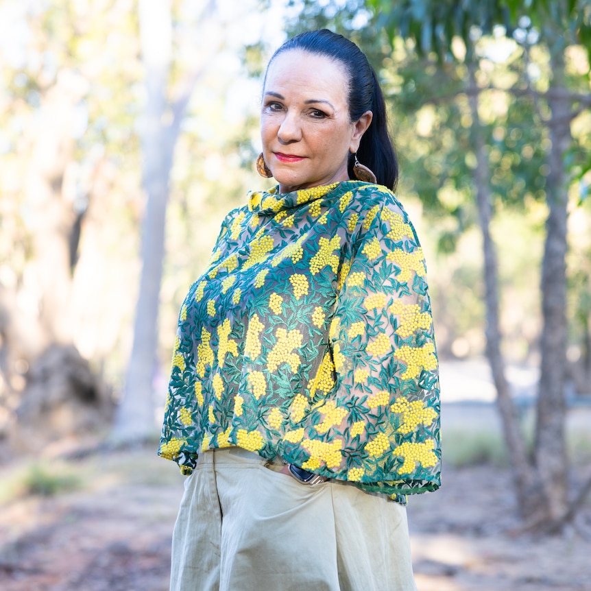 an indigenous woman with a black ponytail wears a top with a green and yellow wattle design, the bush in the background