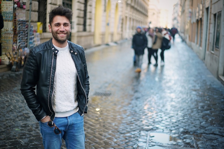 A young man wearing a leather jacket smiles in an Italian street.