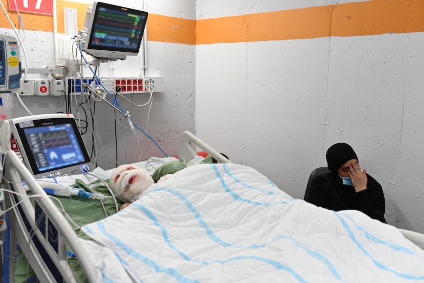 A small child wrapped up in bandages lies in a hospital bed while a woman rests her head in her hand