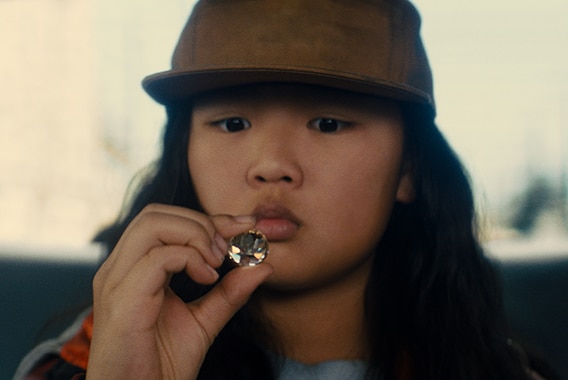 A young girl wears brown flat brim baseball cap and sits in back seat of vehicle and examines large diamond.