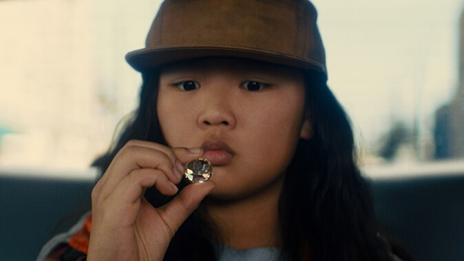A young girl wears brown flat brim baseball cap and sits in back seat of vehicle and examines large diamond.