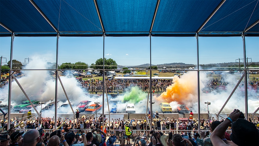 Crowds look onto dozens of cars doing a collective burnout, with colourful smoke towering into the air.