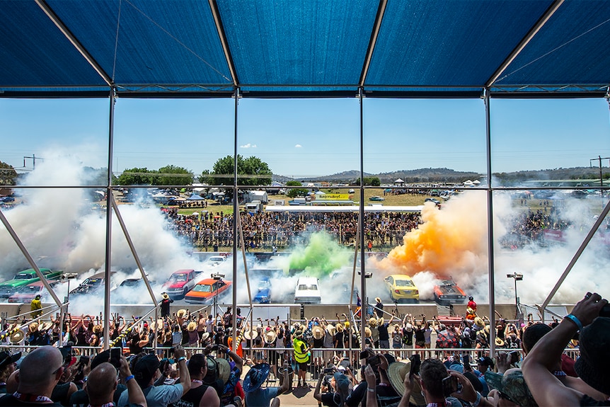 Crowds look onto dozens of cars doing a collective burnout, with colourful smoke towering into the air.