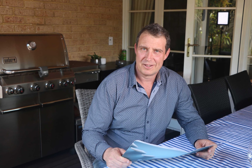 A man sits at an outdoor table holding some papers with a barbecue in the background.