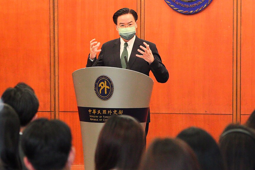 A man in a suit and face mask gestures with both hands as he speaks to media from behind a lecturn.