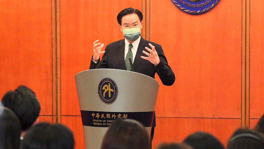 A man in a suit and face mask gestures with both hands as he speaks to media from behind a lecturn.