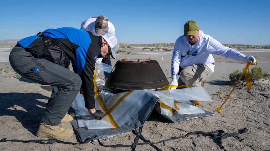 Three people in blue and white clothing place a dark grey capsule on a sheet of plastic in the middle of the desert.
