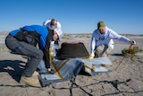 Three people in blue and white clothing place a dark grey capsule on a sheet of plastic in the middle of the desert.