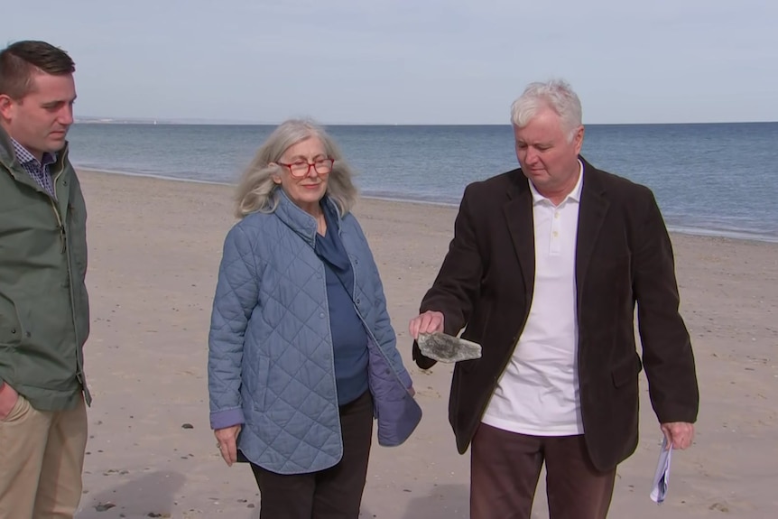 Two men and a woman examine a slate of asbestos while they're on the beach