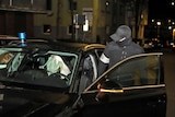 A German police officer in a cap and a mask gets into a car with a person with a white bag over his head in the backseat.