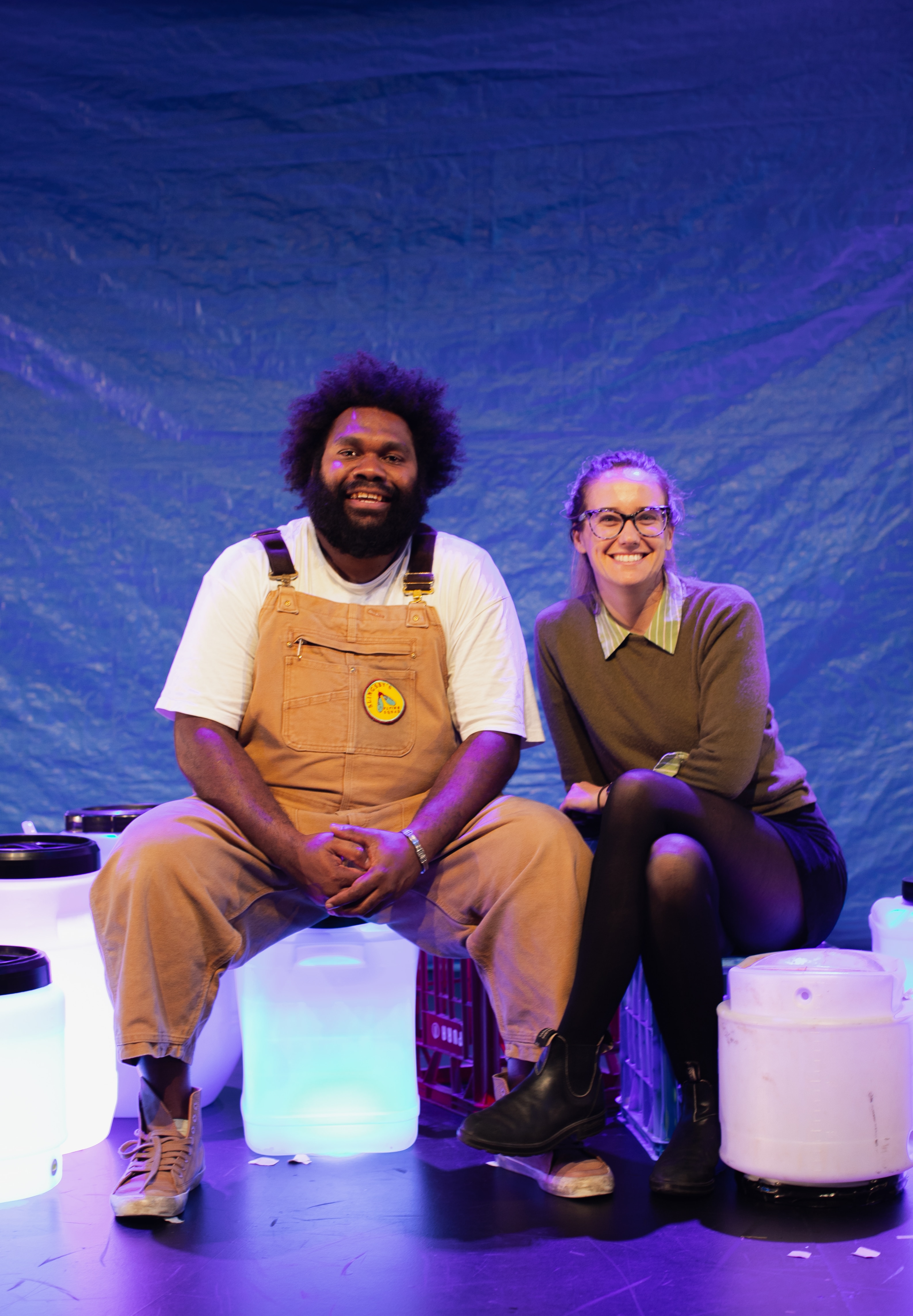 Indigenous man wearing caramel overalls sits on a blue-lit stage beside a white woman in khaki jumper with glasses.