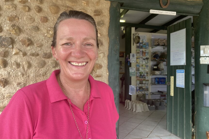 A woman smiles, she is wearing a pink polo shirt. There is a building with travel brochures in the background.
