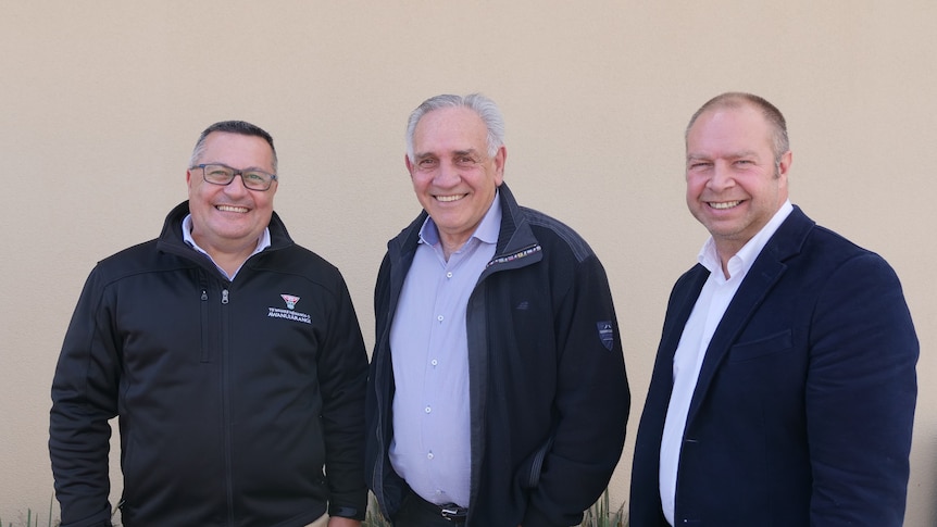 Three men stand side-by-side, all wearing black or navy coats, smiling at the camera. 