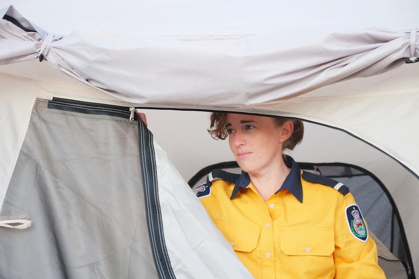 A woman zips up a tent, wears a yellow uniform with badges and lapels on the sleeves, slight smile.