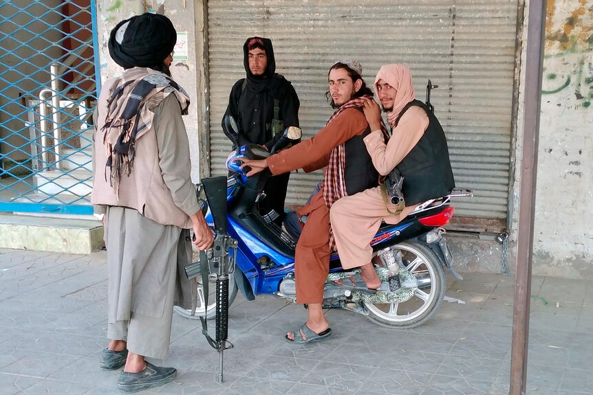 Two men sit on a motorbike, while another talks to them and leans on a weapon. 