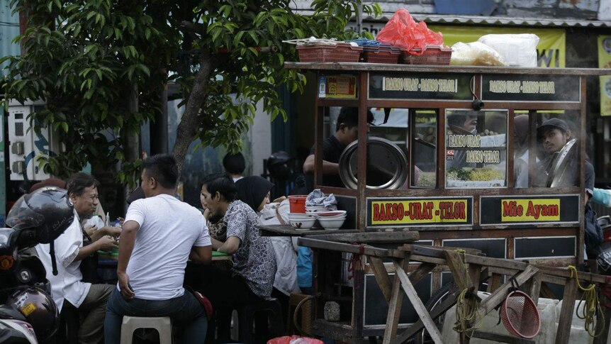 A street-vendor cart featuring signs that read 'Bakso' and 'mie ayam' surrounded by customers eating at plastic stools.