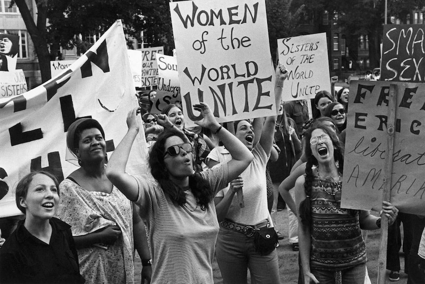 A black and white image shows women holding a sign that says: 'women of the world unite'