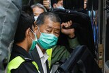 A man, surrounded by poice and supporters, enters a prison van, wearing a suit and face mask