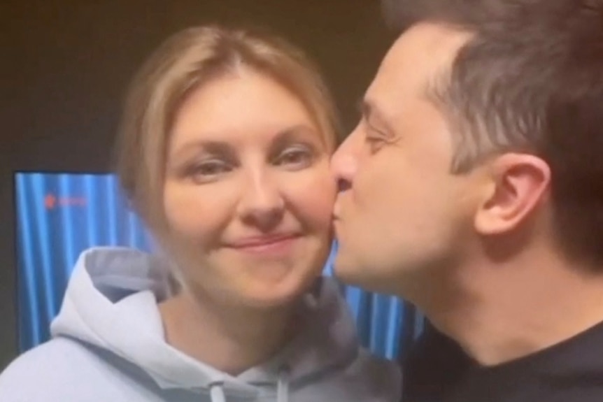 A woman smiles and holds a bunch of flowers as a man kisses her check