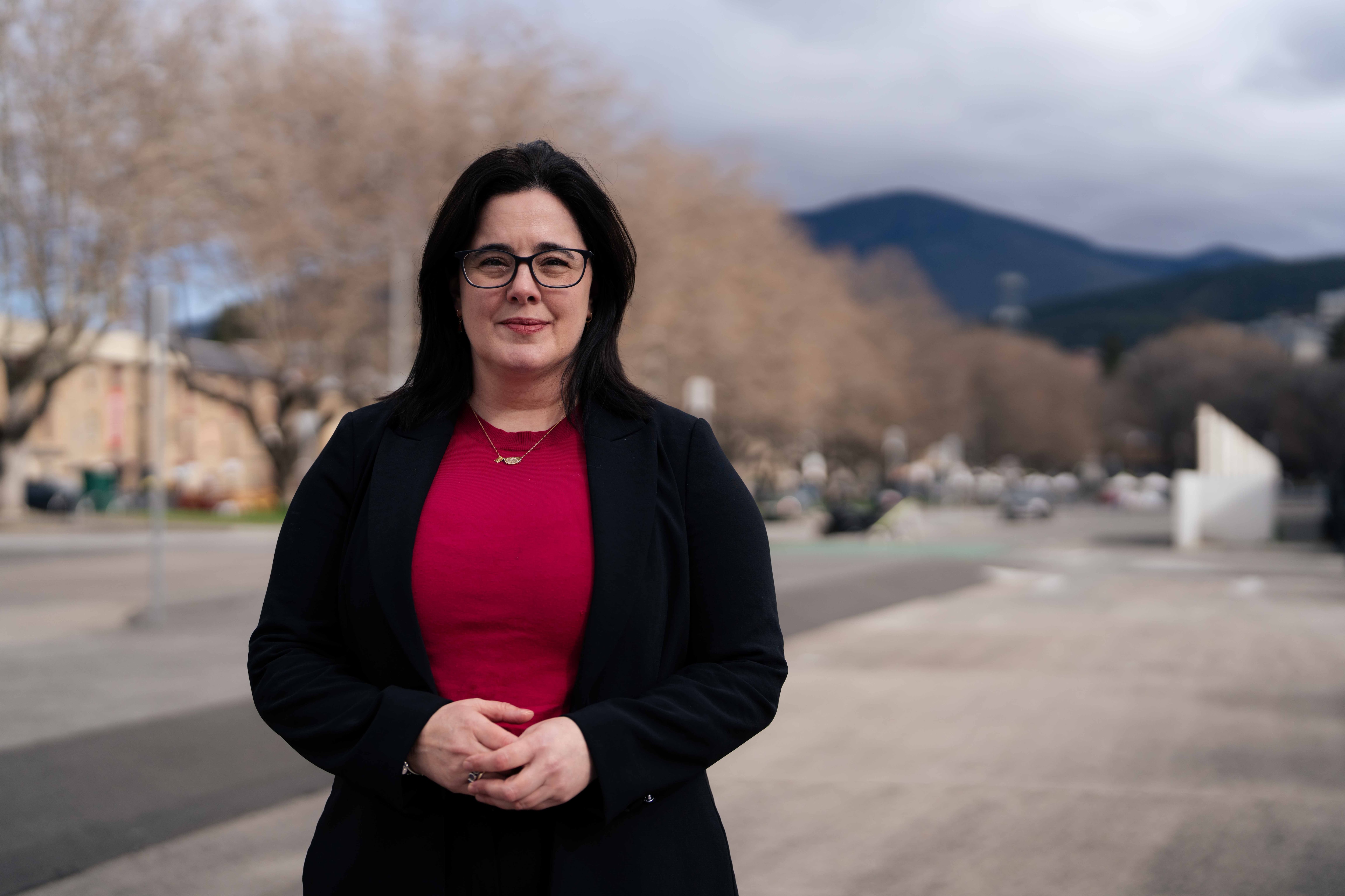 Uma mulher de blusa vermelha e jaqueta preta sorri para uma foto.