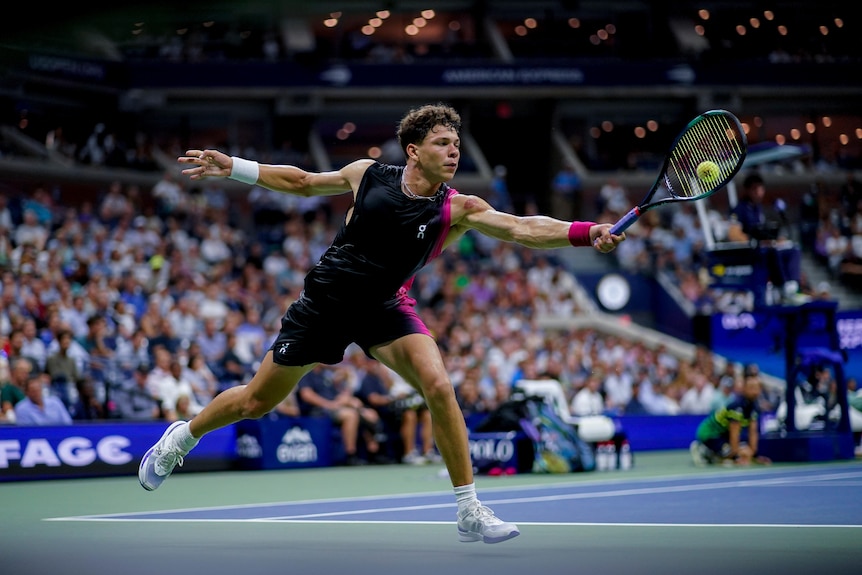 A young man wearing black shorts and singlet with a little pink reaching out with a tennis racket to hit a ball