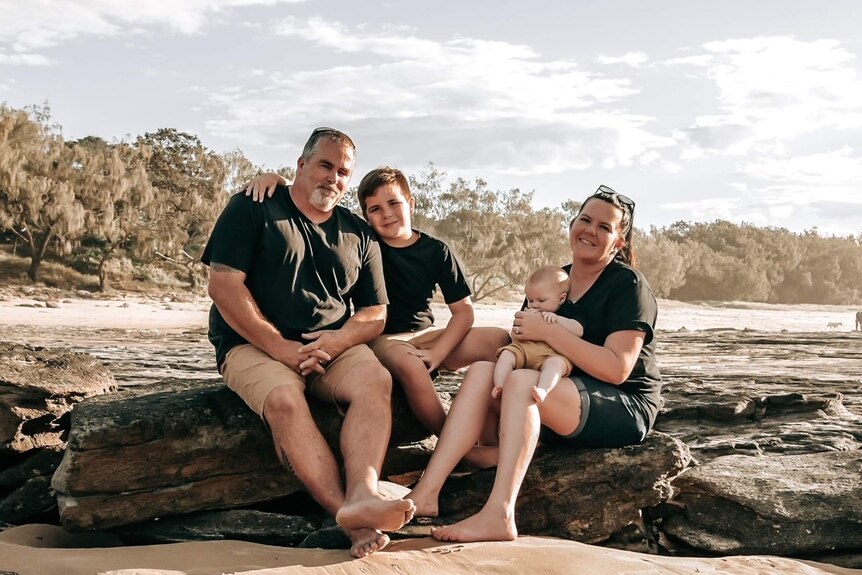 Family of mum, dad, young boy and baby sitting on rocks at the beach, all smiling