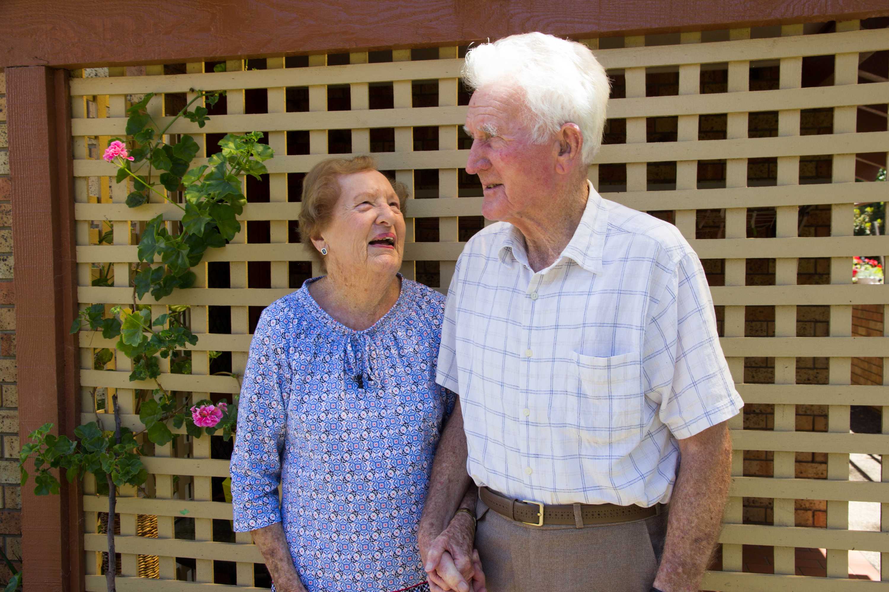 An elderly woman looks up adoringly at an elderly man she is holding hands with.