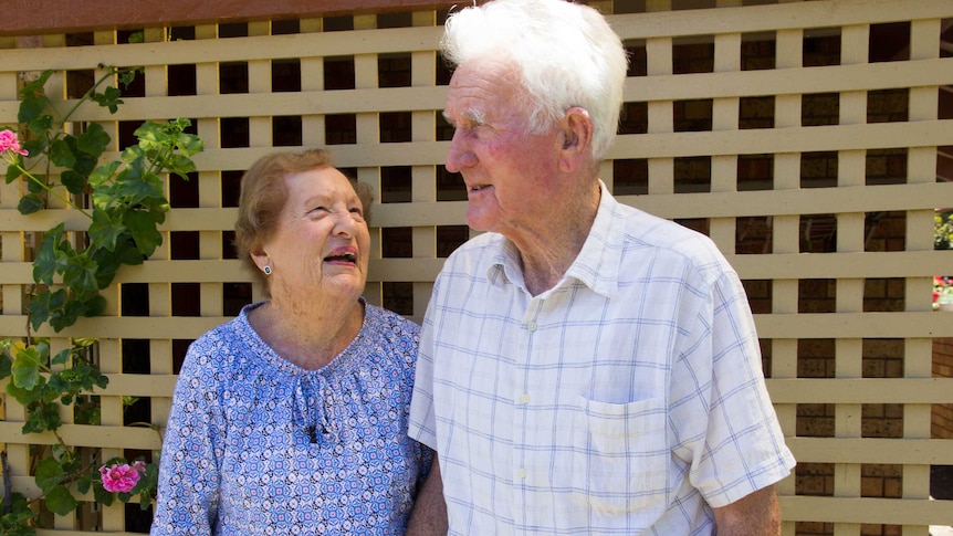 An elderly woman looks up adoringly at an elderly man she is holding hands with.