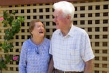 An elderly woman looks up adoringly at an elderly man she is holding hands with.