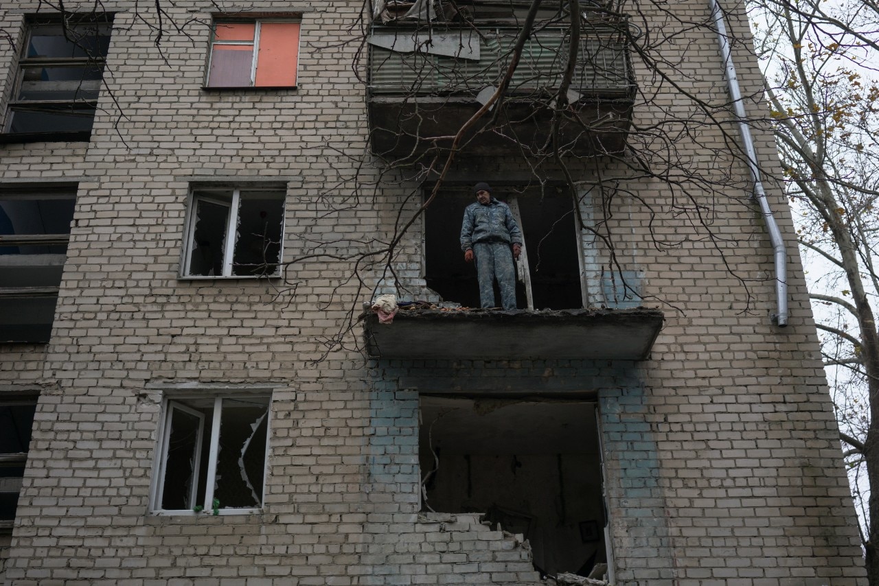 A man surveys the damage to an apartment, through a large hole in the wall, standing on what remains of a balcony.